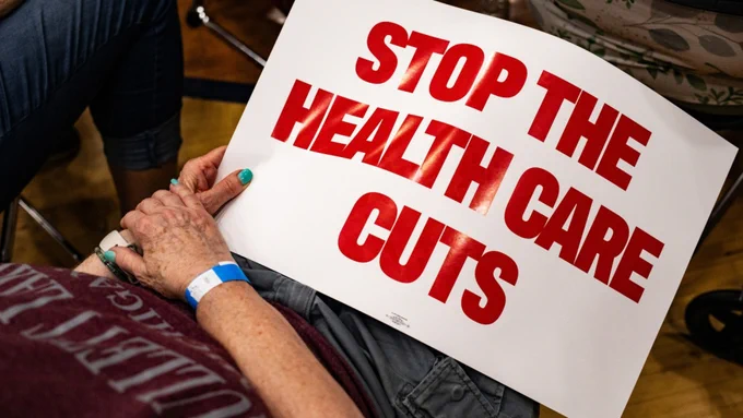 A person is holding a sign that reads, "Stop the health care cuts" in bold red letters.