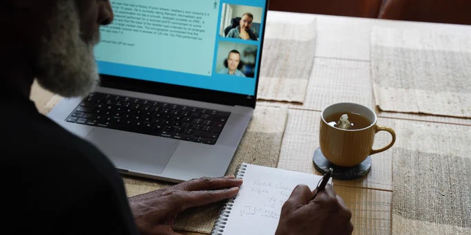A person sits at a table with an open laptop displaying a video call and a document. They write notes in a notebook. A cup with a hot beverage is nearby.