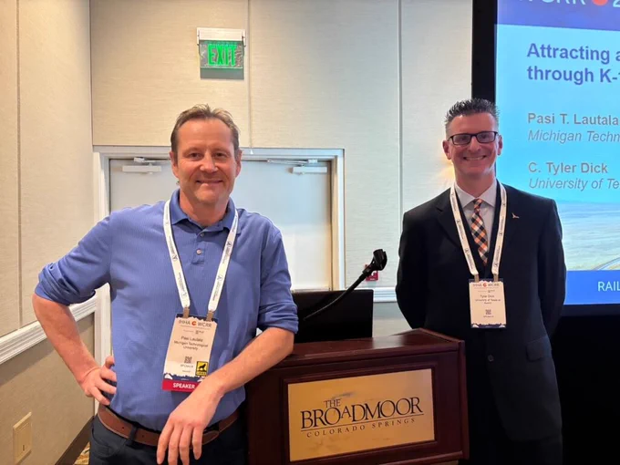 Dr. Pasi Lautala and Dr. C. Tyler Dick stand beside a podium at the World Congress on Railway Research conference in Colorado Springs, Colorado. Lautala is wearing a blue polo shirt and conference badge, while Dick is wearing a dark suit and patterned tie with a conference badge. A presentation slide behind them displays the title of their award-winning paper about attracting a new generation of railroad professionals through K-12 activities and programs.