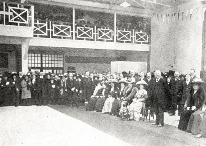 A formal gathering of men and women in early 20th-century attire inside a large hall with a balcony.