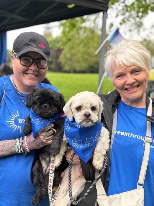 Two people holding two small dogs wearing blue bandanas at an outdoor community event.