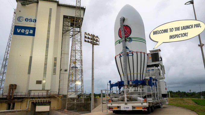 A photo showing a lorry driving along a narrow road with a large rocket nose cone standing upright on its trailer. Ahead of the lorry (to the left in this image) is a tall rectangular cream-coloured building, which has the ESA and Vega logos on its side. The building is surrounded by metal pylons. A speech bubble reads 'Welcome to the launchpad, Inspector!'.