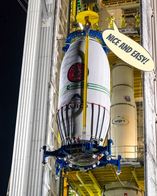 A photo showing the nose cone of a rocket hanging from a crane in front of an open technical building. Inside the building stands the rest of the rocket, with a platform at the top where people stand, waiting for the nose cone to arrive. A speech bubble reads 'Nice and easy!'.