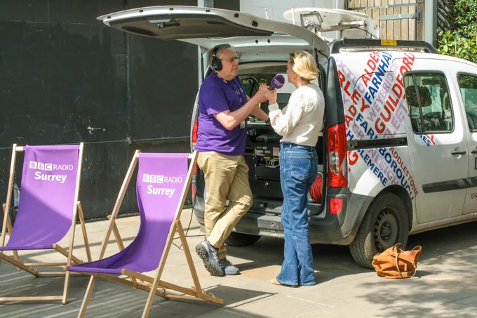 Image shows Commissioner Lisa Townsend in blue jeans and a white top from behind chatting with BBC Surrey radio presenter James Cannon on his stick microphone live on the radio.  They are standing at the back of a radio satellite van which is open showing technical equipment.  In front of them are two purple BBC Radio Surrey deck chairs.