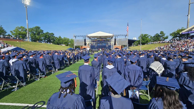 Graduates in blue regalia stand at Paulson Stadium during a commencement ceremony.