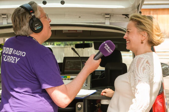 Image shows a mid shot in profile of Commissioner Lisa Townsend in a white top chatting with BBC Surrey radio presenter James Cannon on his stick microphone live on the radio.  James Cannon is wearing a purple branded Radio Surrey tee-shirt They are standing at the back of a radio satellite van which is open showing technical equipment.  Lisa is smiling broadly.