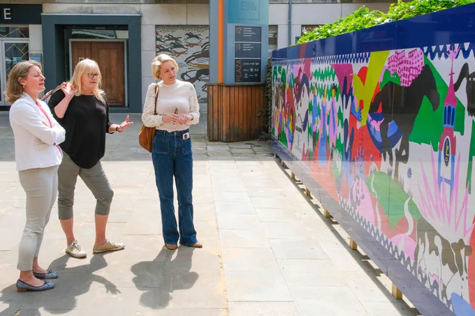Image shows Lisa Townsend who is wearing a white top and blue jeans looking at a vibrantly coloured wall mural that is depicting the history of Epsom.  She is standing beside a fair haired lady with glasses who is from the Epsom Bid team and also Helen Maguire MP who is wearing a white blazer and cropped stone coloured capri pants.  The three ladies are looking together at the colourful mural