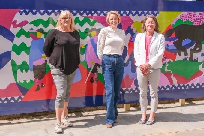Image shows Lisa Townsend who is wearing a white top and blue jeans standing beside a fair haired lady with glasses who is from the Epsom Bid team and also Helen Maguire MP who is wearing a white blazer and cropped stone coloured capri pants.  All three ladies are standing in front of the new vibrantly coloured wall mural in a posed fashion in Epsom Square and are smiling broadly to camera.