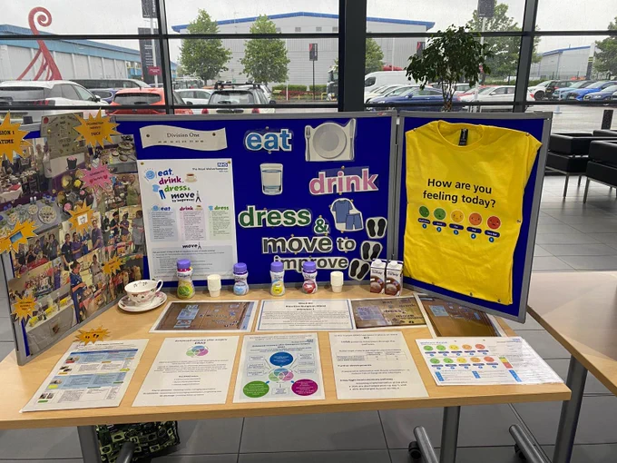 NHS ‘Eat, Drink, Dress & Move to Improve’ exhibition stand showcasing nutrition, wellbeing and Enhanced Recovery After Surgery resources, with promotional posters, a yellow feedback T-shirt and Fortisip drinks on display.