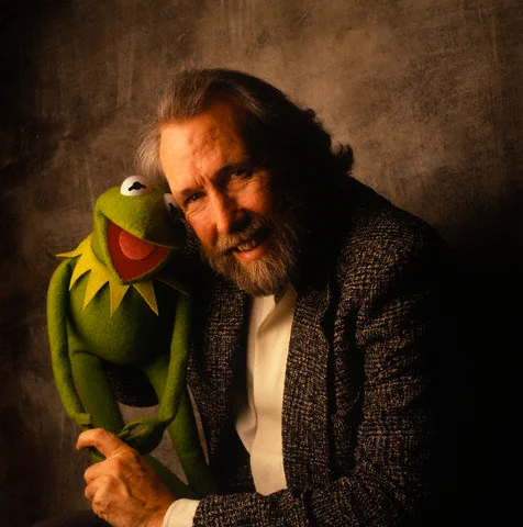 Jim Henson, wearing a speckled dark gray jacket and white collared shirt, smiles as he holds up Kermit the Frog in front of a dark gray background. (Image courtesy of Muppet Wiki)