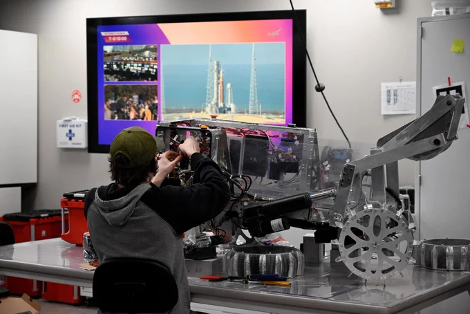 Michigan Tech engineer working on a robotic rover prototype in a lab with a rocket launch displayed on a screen behind.