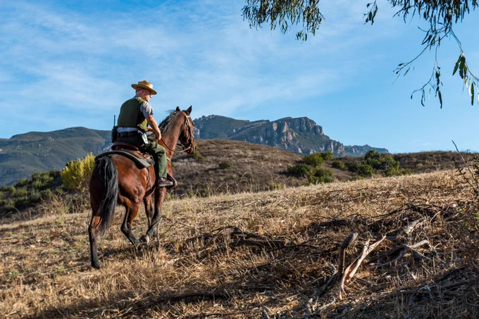 A mounted law enforcement ranger rides a brown horse across dry grassland in a mountainous landscape under a blue sky. The ranger wears a wide-brimmed hat and carries gear while patrolling public lands in a remote area.