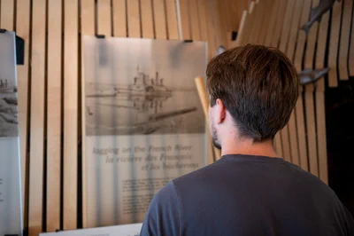 A person is standing in front of an exhibit, looking at historical information about the French River. The display includes text and images related to the river's history. The setting has wooden wall elements.