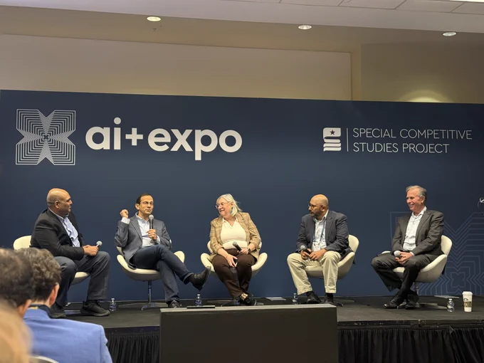 A group of five people sit on a stage at a panel discussion. The backdrop features the logos for "AI   Expo" and "Special Competitive Studies Project." The individuals are engaged in conversation, seated in chairs across the stage.