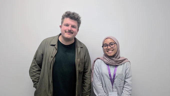 An image of two people standing side-by-side against a plain white background. On the left, a man with light-colored spiky hair and a mustache is wearing a dark t-shirt and an olive-green open jacket. On the right, a smiling woman wearing glasses and a mauve hijab is dressed in a grey button-down shirt with purple lanyards around her neck.