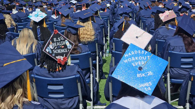 Graduates sitting at commencement. One grad cap says, "Hardest workout ever! BSK '26, James 1:4" and another says, "B.S. Health Science."