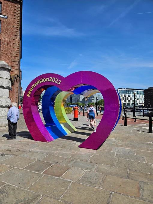 3 large half hearts embedded into a pavement. Each heart has a different colour – pink, blue and yellow. The words #eurovision2023 feature at the top of the first heart. Red post box in the distance, blue sky and red brick dock building to the left hand side.