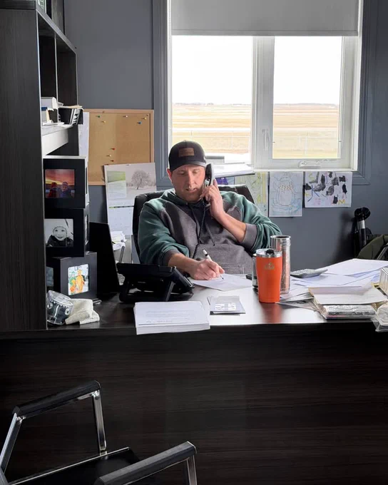 Man sitting in an office, writing notes and talking on a telephone. His hat displays a logo for Double Diamond Farm Supply.