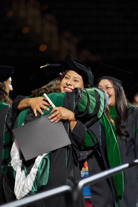 Graduate in a black gown and cap embraces another person on stage while holding a diploma cover; others in academic regalia stand nearby under stage lighting.\n
