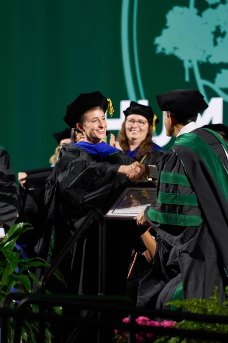Graduate in cap and gown shakes hands with a faculty member while receiving a diploma on stage; additional faculty and graduates are visible in the background.