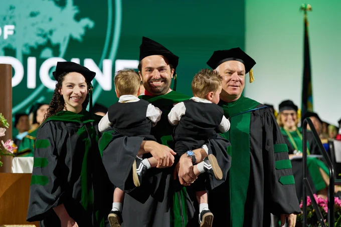 Graduate in a black cap and gown posing on stage with two young children being held; a green backdrop and other ceremony participants appear behind them.