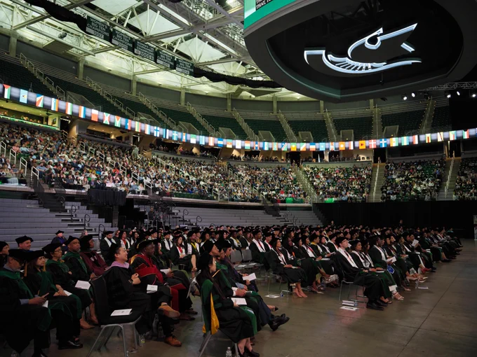 Large indoor commencement ceremony in a stadium-style arena with rows of graduates in caps and gowns seated on the floor, a full audience in tiered seating, and a ring of international flags displayed around the venue beneath championship banners and overhead lighting.\n
