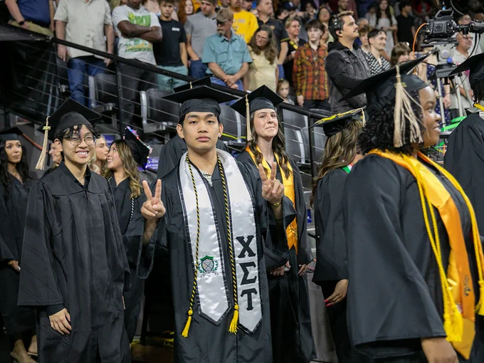 A group of graduates in caps and gowns stand together indoors. One person in the center flashes a peace sign.