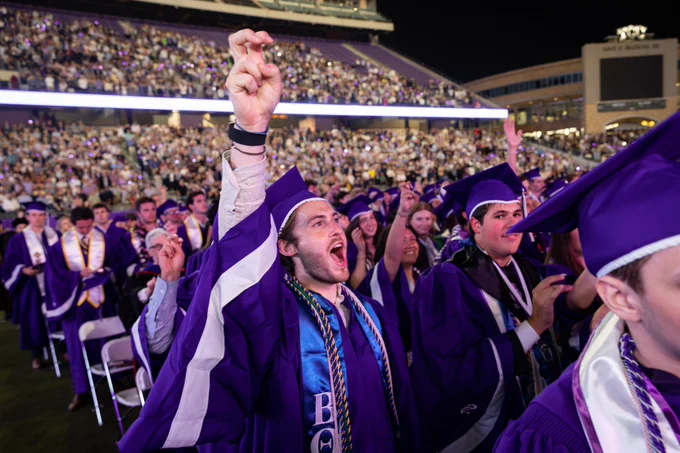 Graduates in purple robes and caps celebrate at a commencement ceremony in a packed stadium. One person in the foreground raises a hand in excitement. The crowd in the background is filled with people, many with phones raised, capturing the moment.