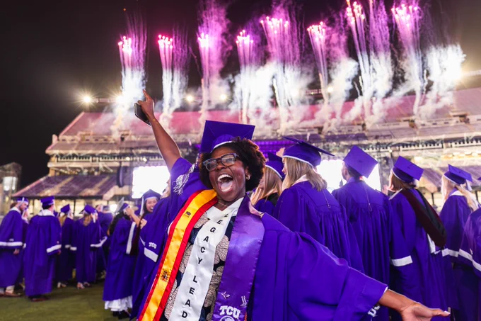 A person wearing a purple graduation gown and cap, decorated with a "Legacy Leaders" sash, is cheering and posing energetically. They are surrounded by other graduates in similar attire. In the background, purple and white fireworks light up the night sky above a large, illuminated stadium, identified as TCU (Texas Christian University).