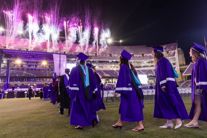 Graduates in purple robes and caps walk in line on a football field during a night ceremony. Fireworks light up the sky in the background. Stadium seating is visible.
