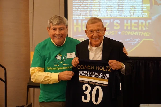 Two men are standing in a room, one wearing a green shirt with a logo and the other in a black suit, holding a framed jersey with the number 30 and the name 'Coach Holtz' on it.