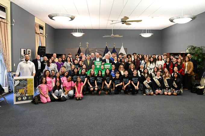 A large group of people, including women and men of various ages, pose for a group photo in a spacious room. Some are holding signs and banners. A few flags are visible in the background, adding a formal atmosphere.