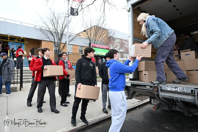 A group of people, including several young adults, are standing outside a building, passing boxes to each other and loading them into the back of a truck. It appears to be a community effort or volunteer activity.