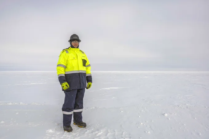 A person stands on a vast snowy landscape wearing a bright yellow jacket, dark pants, a helmet, and gloves.