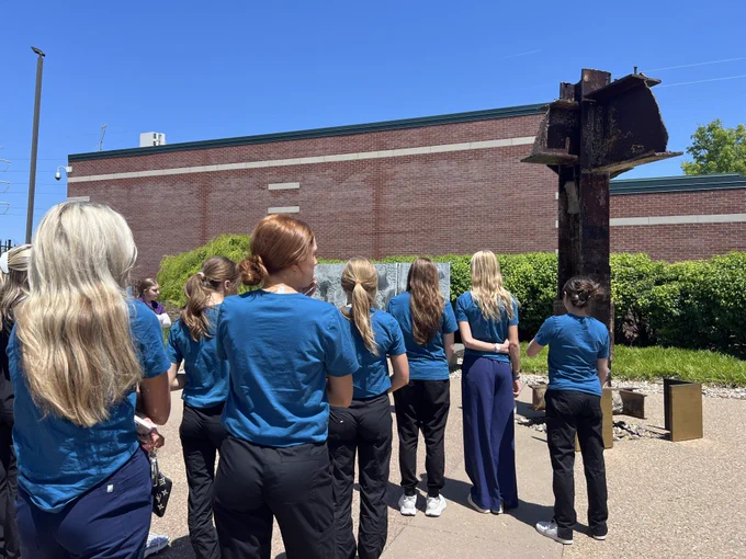 A group of young people in blue shirts standing in front of a large brick building (OP Fire Dept.), looking at a 9/11 memorial.