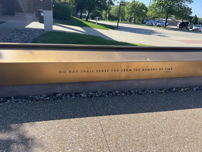 A concrete wall engraved with the text "NO DAY SHALL ERASE YOU FROM THE MEMORY OF TIME" in a public outdoor setting with trees and a road visible.