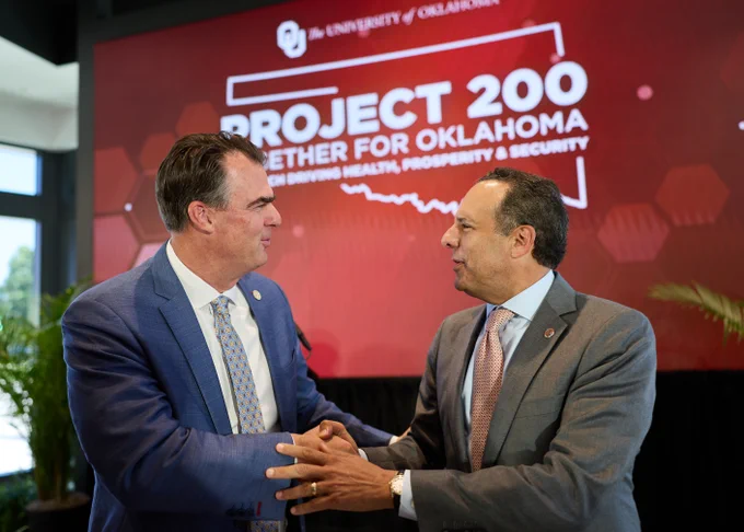 University of Oklahoma President Joseph Harroz Jr. and Oklahoma Gov. Kevin Stitt shake hands and speak during a Project 200 event. Behind them, a large screen displays the OU logo and the words “Project 200: Together for Oklahoma,” focused on driving health, prosperity and security initiatives across the state. Both men are dressed in business attire and standing in front of a red presentation backdrop.