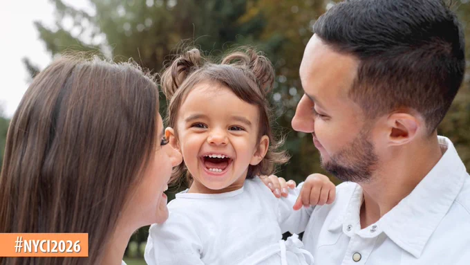 A young family outdoors with parents holding their toddler between them in a park setting.