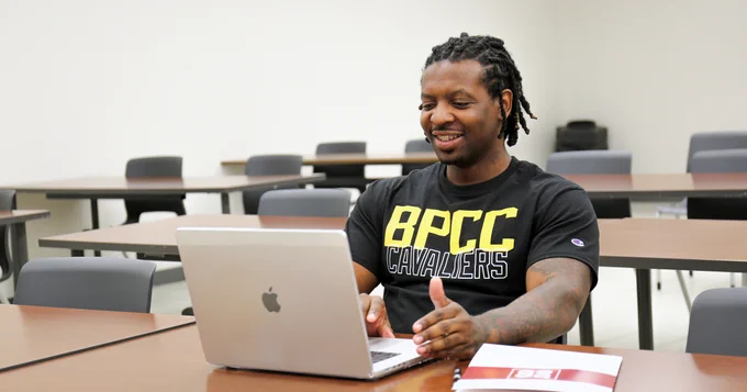 A person is sitting in a classroom, smiling and using a MacBook. They are wearing a shirt with "BPCC Cavaliers" printed on it. Nearby is a BPCC branded folder on the table.