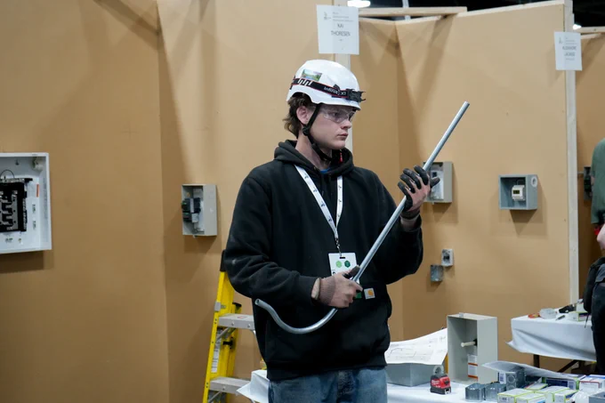 A person stands in a workshop area holding a metal pipe. They are wearing a safety helmet and goggles. The background shows various electrical boxes and tools on tables.