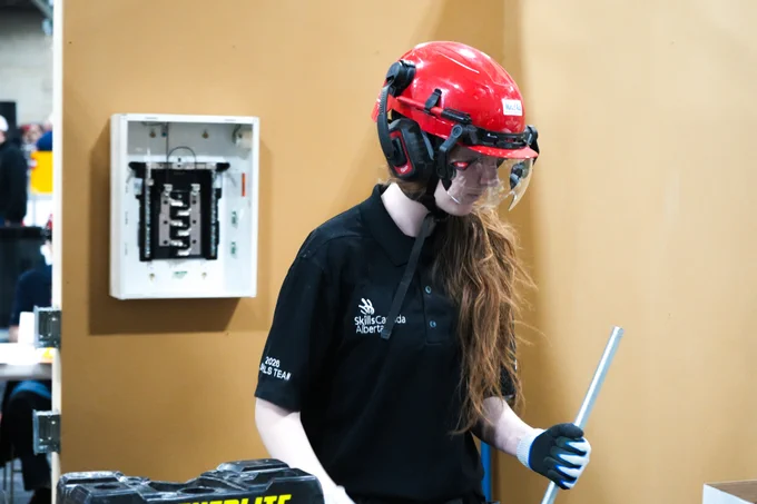 A person with long hair wearing a red helmet and protective gear participates in a Skills Canada Alberta competition. They are holding a metal rod, and there is an electrical panel on the wall behind them.
