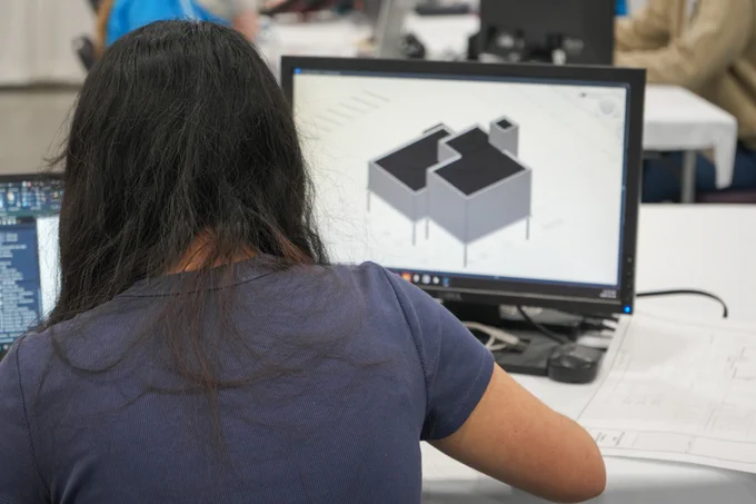 A person is working at a computer, viewing a 3D architectural design on the screen. The design appears to be a building structure. In the background, other individuals are also working at computers.