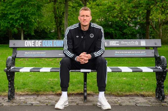 Dan Burn sitting on a themed bench with the words "One of Our Own" and logos of Newcastle United Foundation and Newcastle City Council. The person is wearing a black Adidas tracksuit with white stripes and white sneakers. The background features green trees and grass.