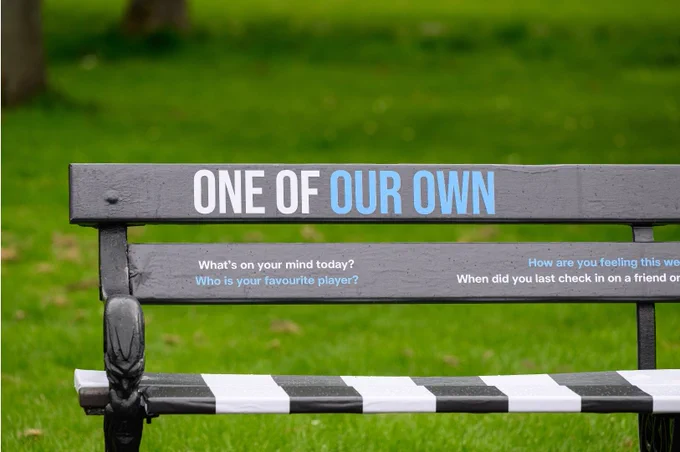 A park bench with "ONE OF OUR OWN" written on the backrest. The slats display reflective questions, including "What's on your mind today?" and "Who is your favourite player?" The bench is situated on a grassy lawn.