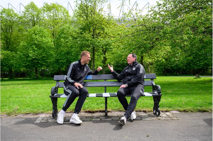 Two people in black athletic wear are sitting and talking on a Newcastle United bench in a park surrounded by lush greenery.