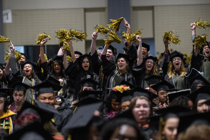 A large group of graduating students cheer in the crowd, waving black and gold pompoms in the air.