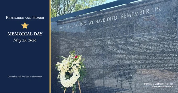 Photo of the Minnesota Vietnam Memorial in Saint Paul, Minnesota. The photo is a closeup of the granite wall with the engraved names of Minnesota's missing and killed in action. Above the names is also engraved "We were young. We have died. Remember us." In front of the wall stands a white floral wreath. To the left of the photo are the words "Remember and Honor, Memorial Day, May 25, 2026. Our offices will be closed in observance."