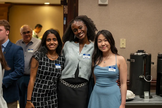 Three smiling young women, \"Urvashi Nagaraj\", \"Sabrina\", and \"Tiffany\" pose closely at an event; each wears a \"HELLO my name is\" tag. A coffee station is visible in the background, with other attendees nearby.