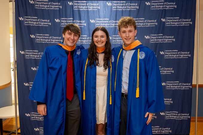 Three smiling graduates pose in front of a University at Buffalo \"Department of Chemical and Biological Engineering\" backdrop. Two men wear blue gowns, one with a red tie. A woman in the center wears a white dress and a blue gown.