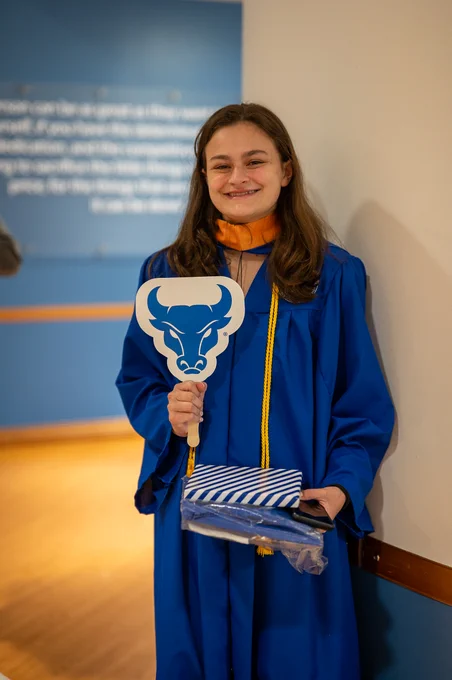 A young woman wearing a blue graduation gown and orange stole smiles, holding a bull logo fan and striped gift. She stands against a two-toned wall, creating a celebratory and academic mood.